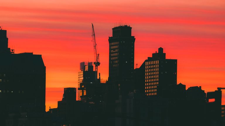 Silhouette of city buildings and a crane against a vivid orange and red sunset sky.
