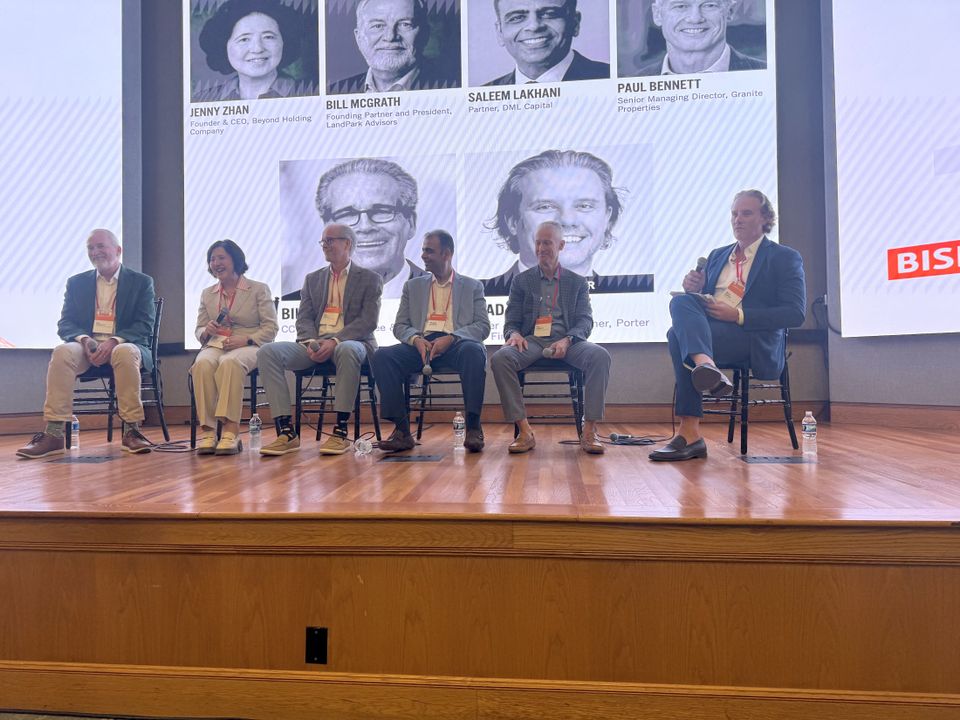 Panel of six people seated on stage with a large screen behind displaying names and photos. They are participating in a discussion or event.