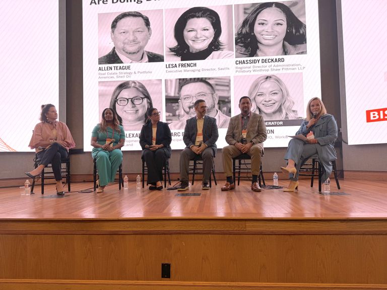 Six people sit on chairs on a stage, participating in a panel discussion. A large screen behind them displays headshots and text.