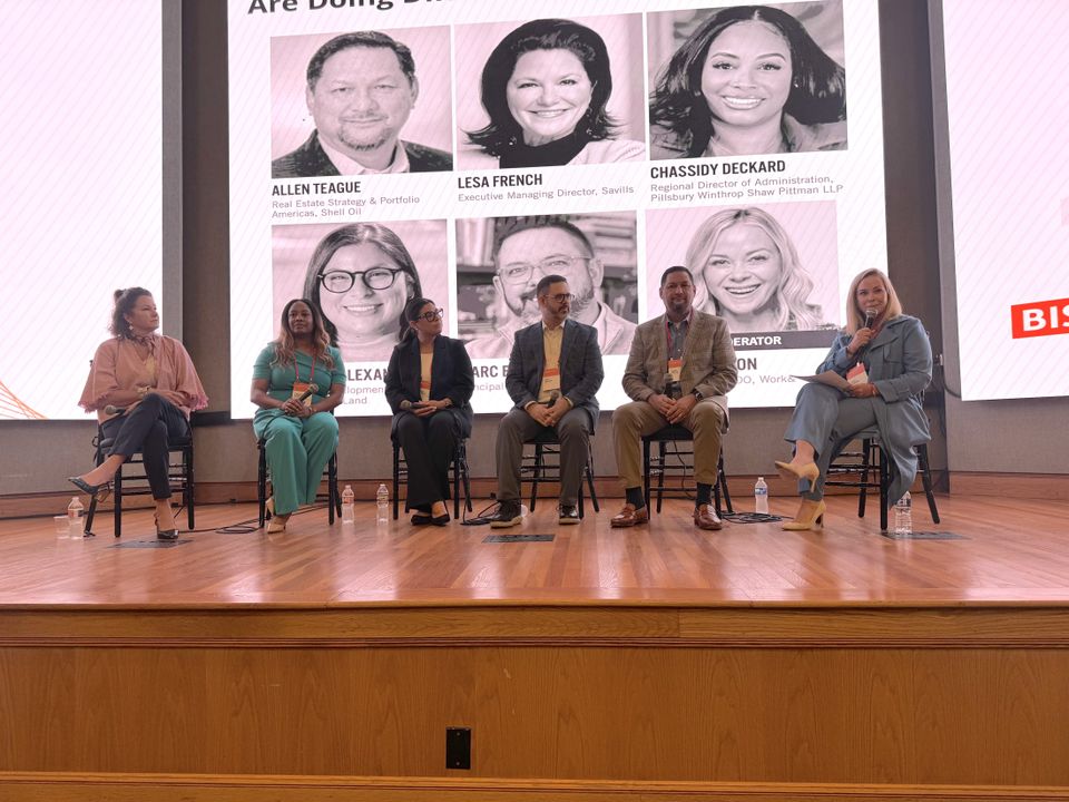 Six people sit on chairs on a stage, participating in a panel discussion. A large screen behind them displays headshots and text.