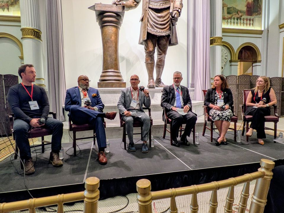 Six people seated on a stage participating in a panel discussion in a formal setting with a large statue in the background.