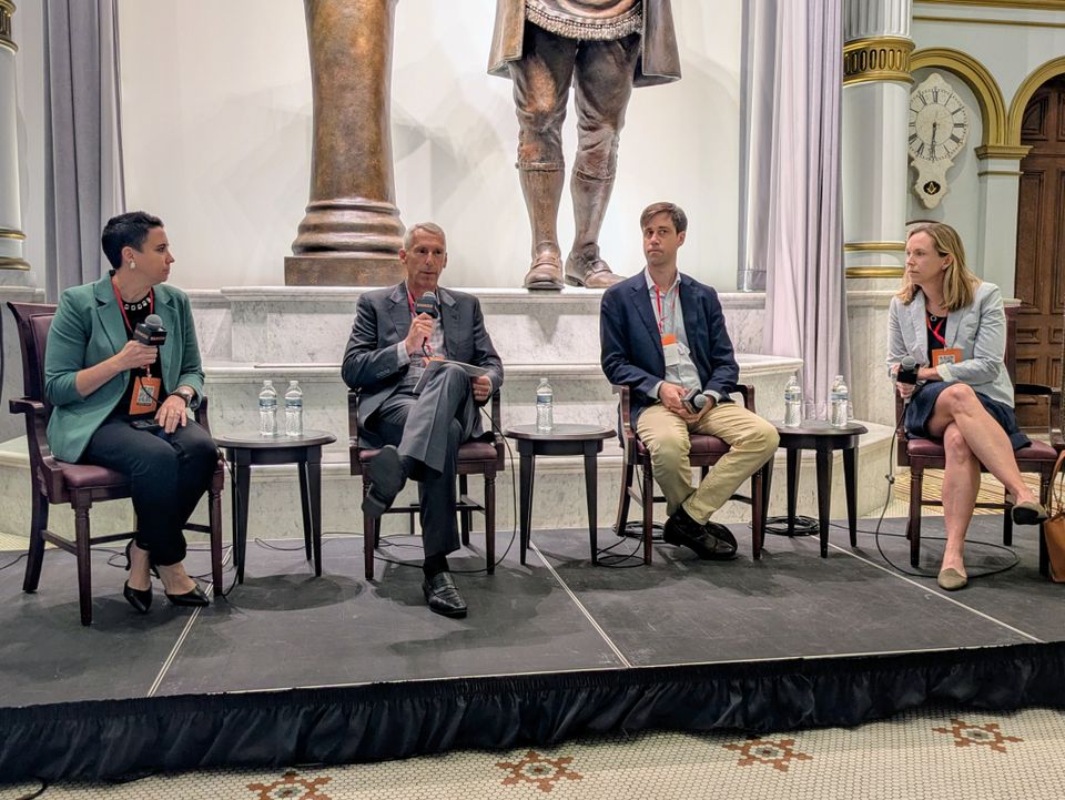 Four people sitting on a stage engaged in a panel discussion, with microphones and water bottles on small tables beside them.