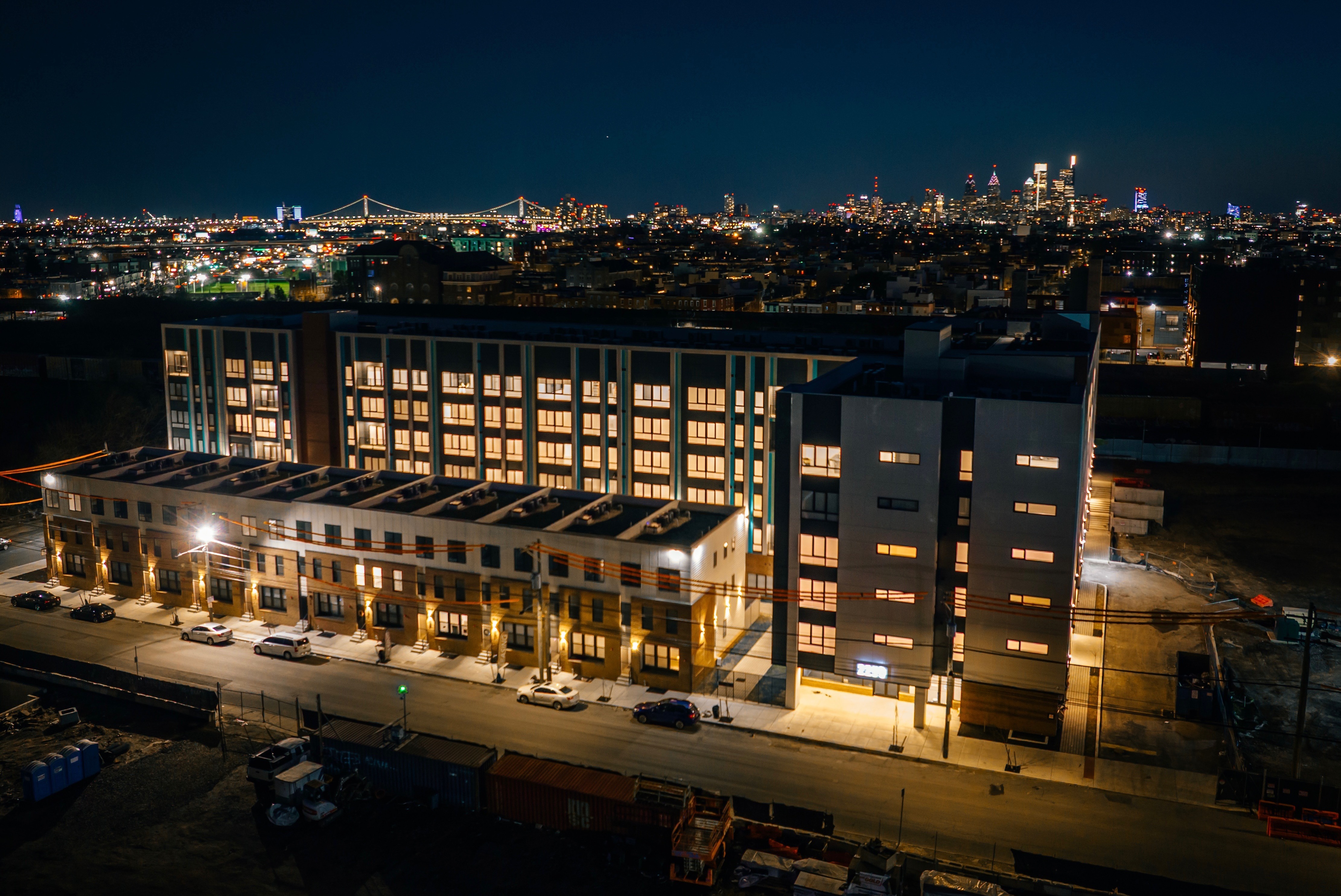 Aerial view of a lit building at night, with a skyline and illuminated bridge in the background.
