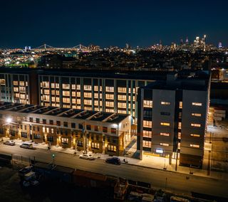 Aerial view of a lit building at night, with a skyline and illuminated bridge in the background.