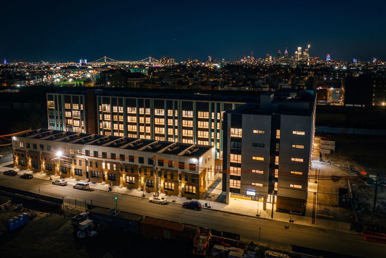 Aerial view of a lit building at night, with a skyline and illuminated bridge in the background.