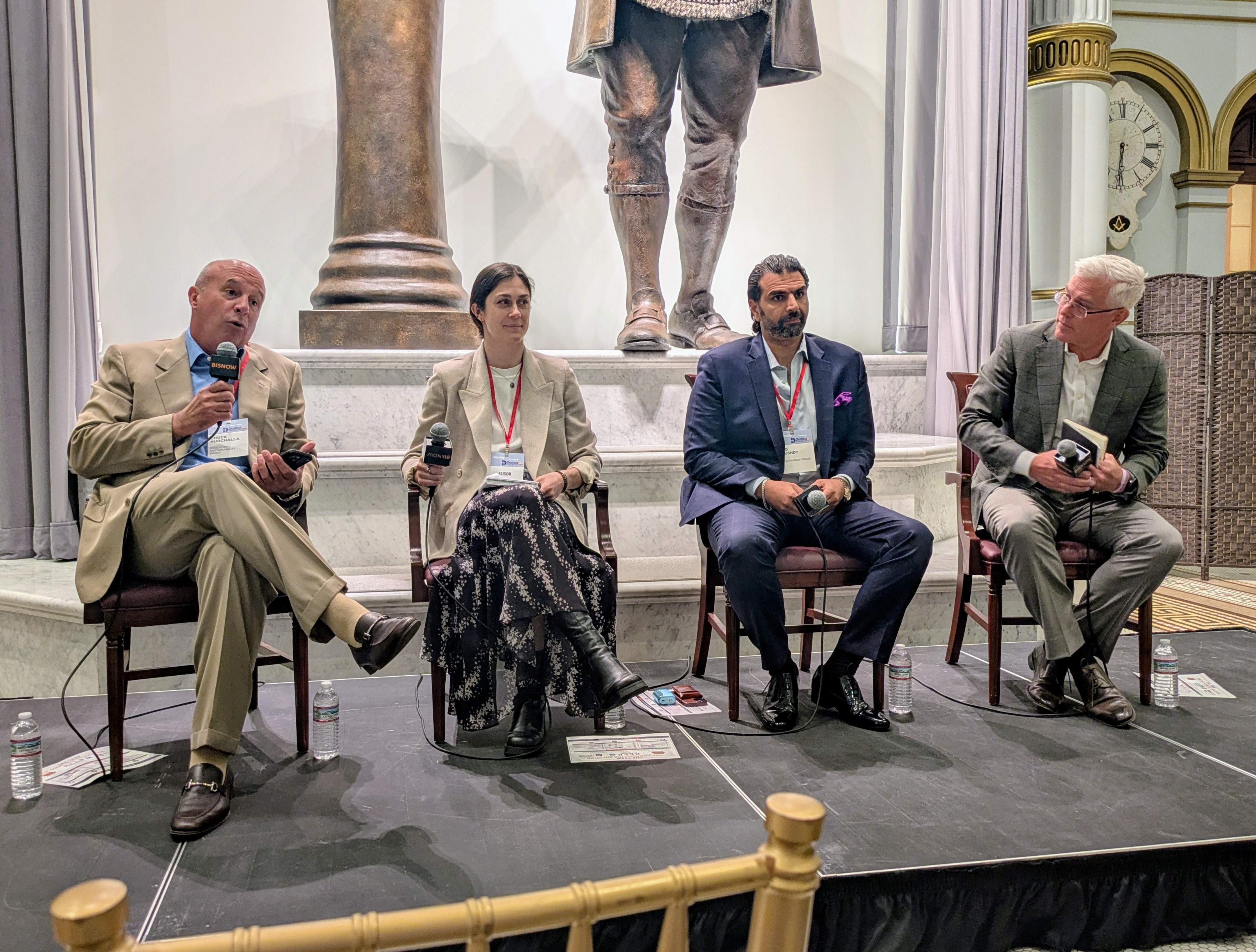 Four people are seated on a stage, each holding microphones, engaged in a panel discussion, with a statue and clock visible in the background.