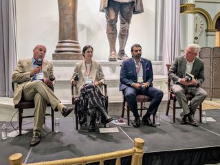 Four people are seated on a stage, each holding microphones, engaged in a panel discussion, with a statue and clock visible in the background.