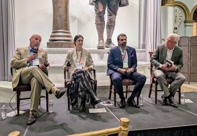 Four people are seated on a stage, each holding microphones, engaged in a panel discussion, with a statue and clock visible in the background.