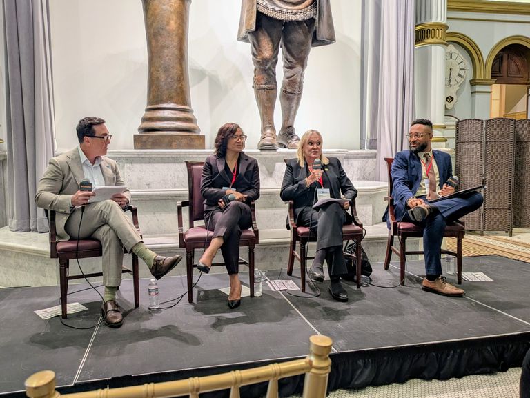 Four people sitting on chairs with microphones, engaged in a panel discussion on a stage with a large statue in the background.