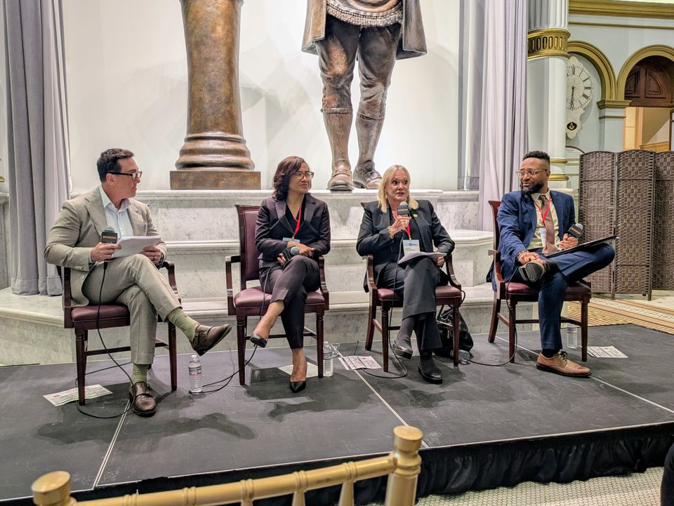 Four people sitting on chairs with microphones, engaged in a panel discussion on a stage with a large statue in the background.