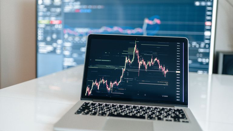 A laptop displays a financial chart with price fluctuations, while a larger screen in the background shows a blurred interface with graphs and data.