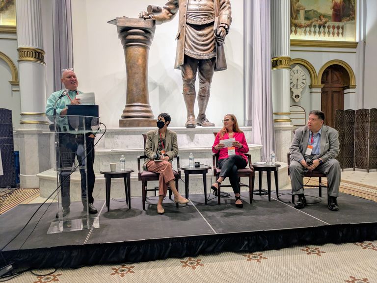 A person speaks at a podium while three others sit on chairs, with a large statue in the background inside an ornate room.