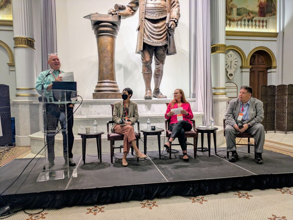 A person speaks at a podium while three others sit on chairs, with a large statue in the background inside an ornate room.