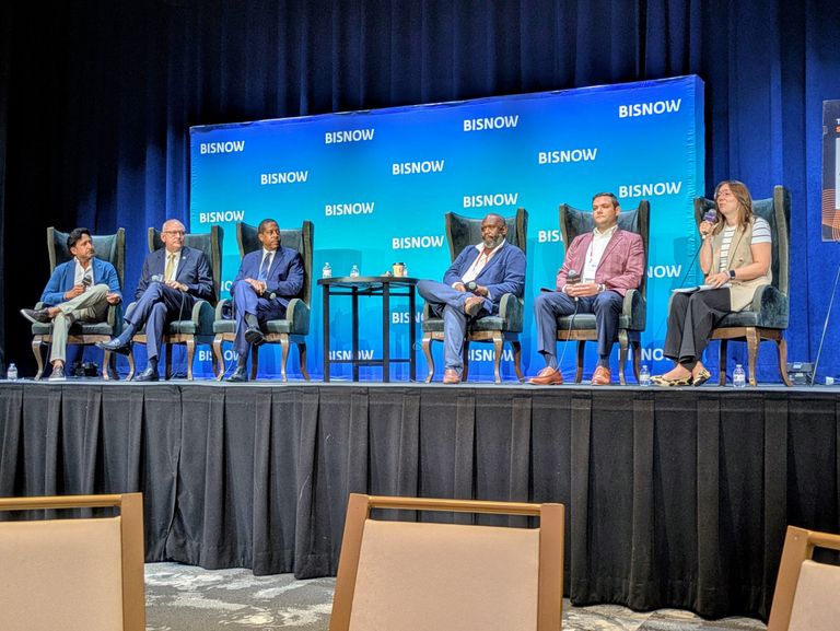 Six people seated on a panel stage with "BISNOW" backdrop, engaged in discussion with microphones, wearing business attire.