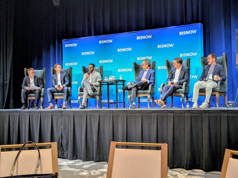 Six men sit on a panel on stage at a BISNOW event, engaging in discussion. The backdrop is blue with BISNOW logos.