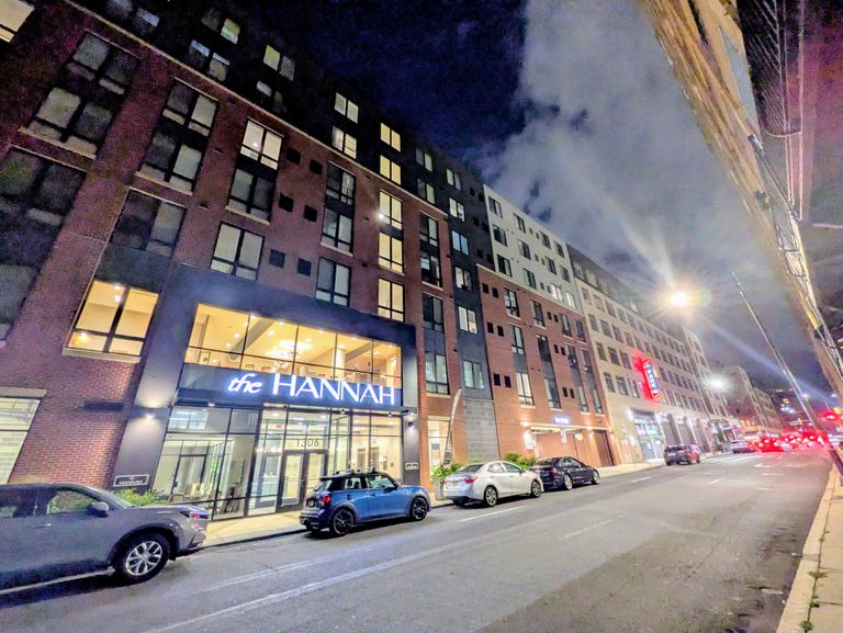 Night street view of a modern multi-story brick building with "The Hannah" sign, lit windows, parked cars, and illuminated streetlights.