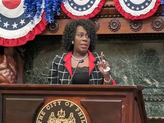 A woman speaks at a podium with the City of Philadelphia seal. The backdrop features patriotic bunting and a carved statue.