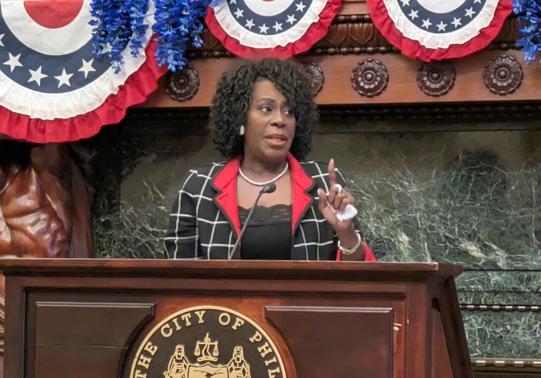 A woman speaks at a podium with the City of Philadelphia seal. The backdrop features patriotic bunting and a carved statue.