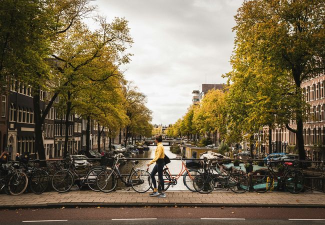 Person in a yellow jacket walking with a bicycle along a canal in Amsterdam, surrounded by autumn trees and parked bicycles.
