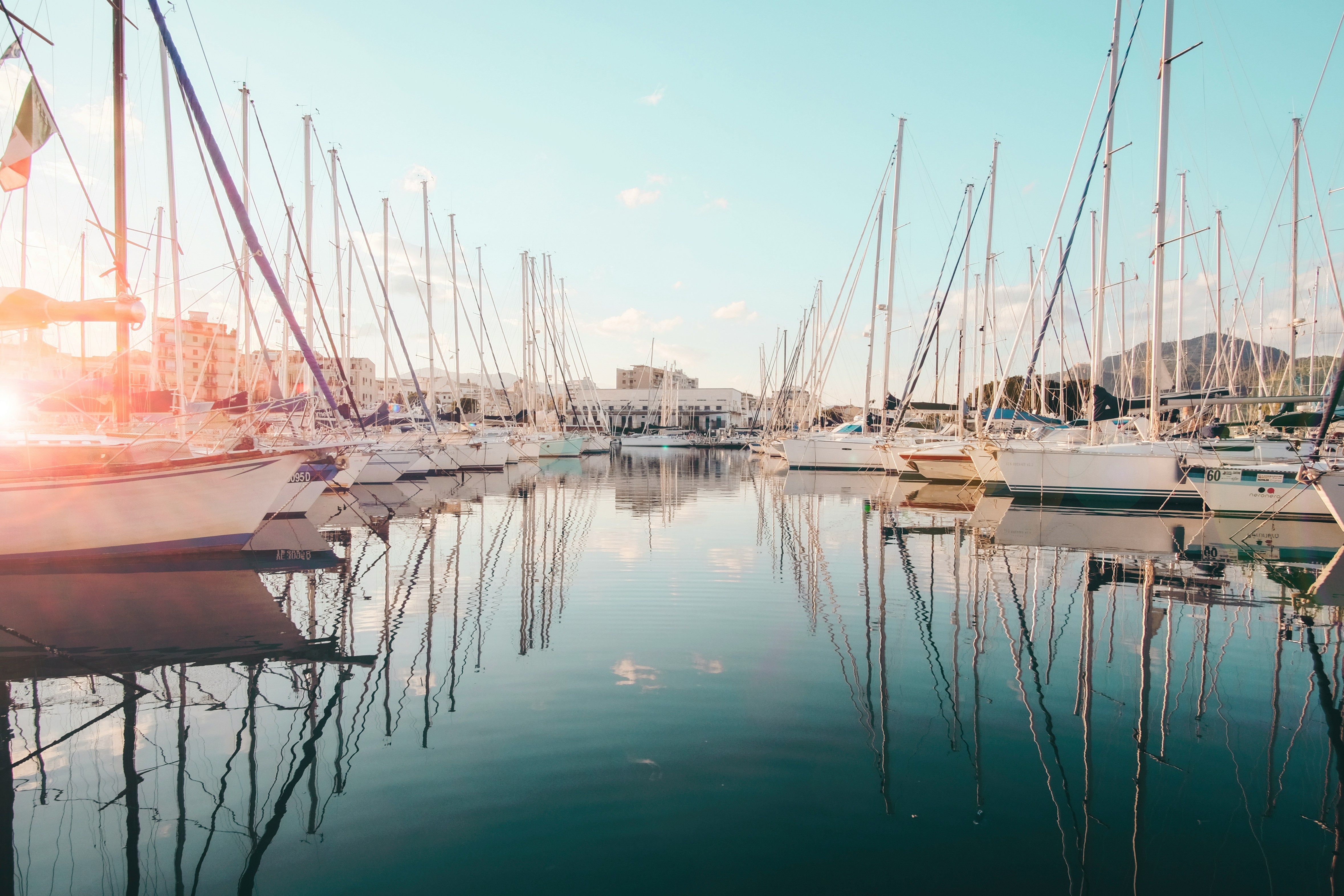 A serene marina with rows of sailboats reflected in calm water, under a clear blue sky with soft clouds and sunlight streaming in.