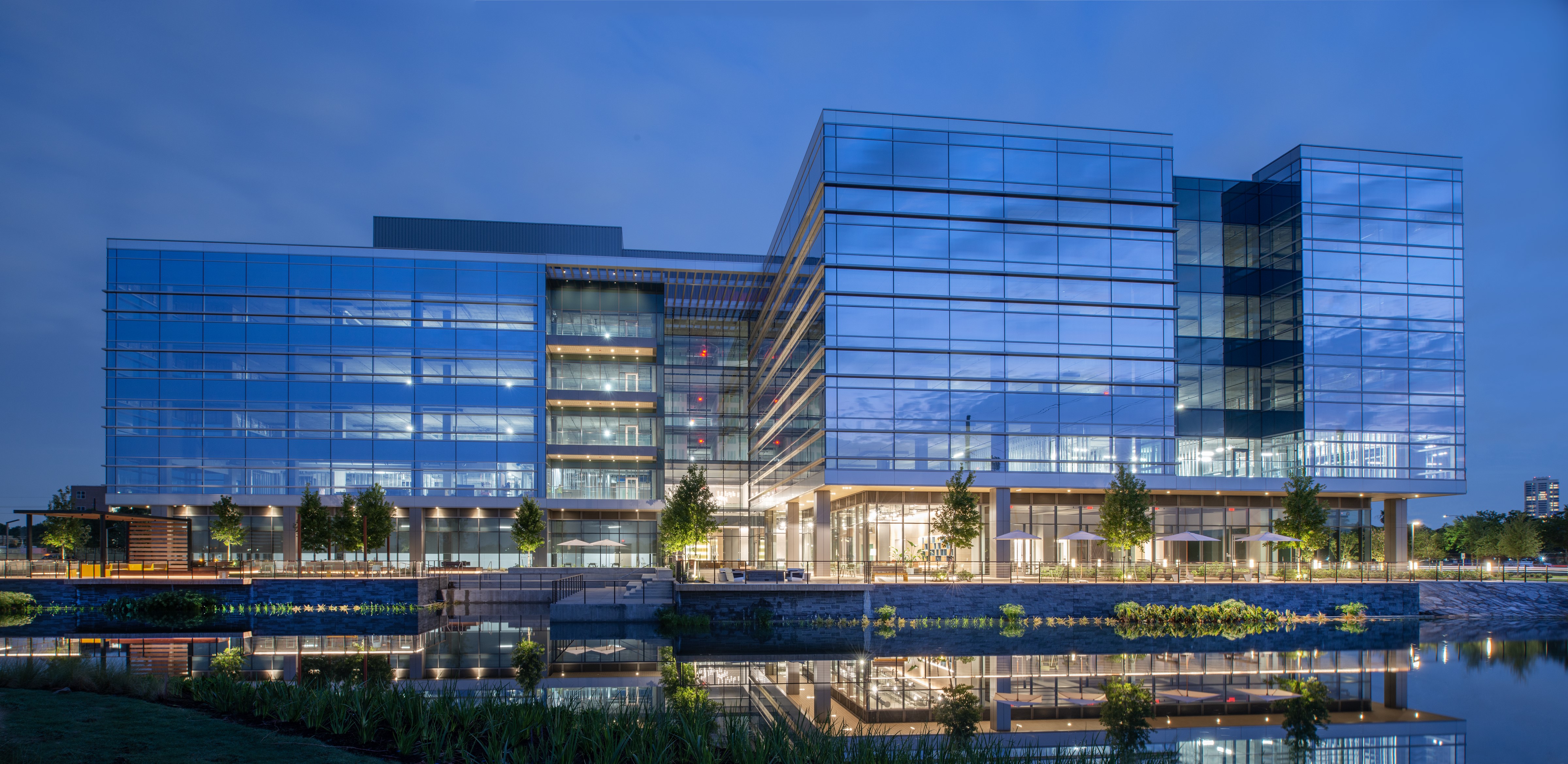 Modern glass building illuminated at night, surrounded by a reflective water body and landscaped greenery.