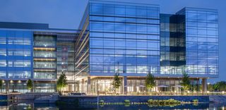 Modern glass building illuminated at night, surrounded by a reflective water body and landscaped greenery.