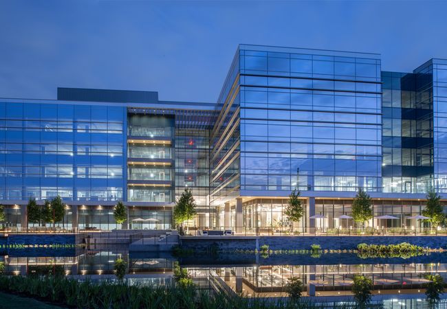 Modern glass building illuminated at night, surrounded by a reflective water body and landscaped greenery.