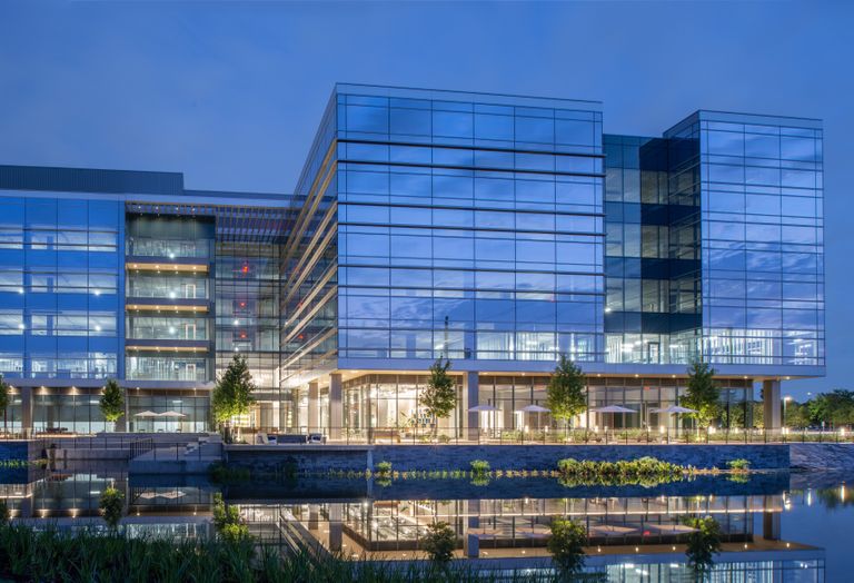 Modern glass building illuminated at night, surrounded by a reflective water body and landscaped greenery.
