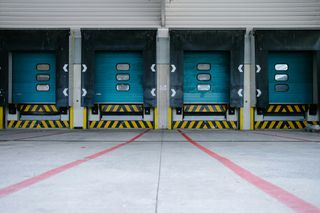 Four industrial loading dock doors with blue panels and protective awnings, marked with red lines and yellow-black safety stripes.