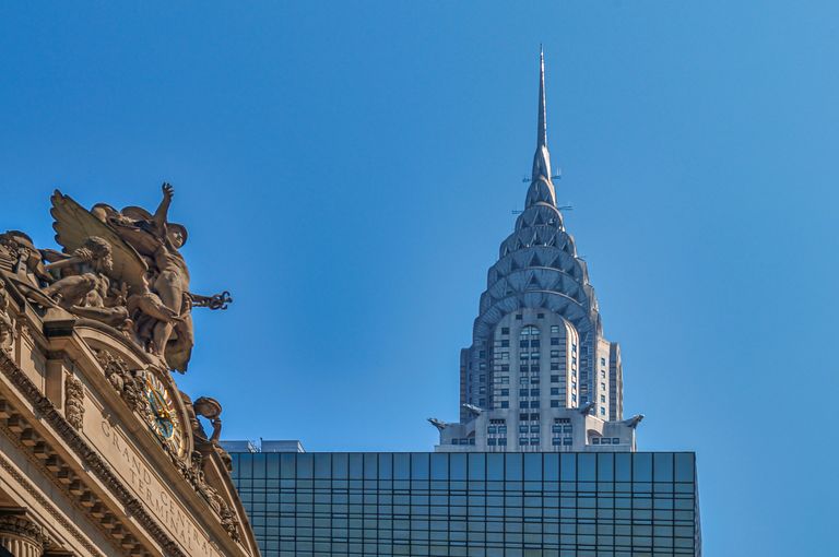 The iconic Chrysler Building spire rises against a clear blue sky, with a decorative architectural detail of Grand Central Terminal in the foreground.