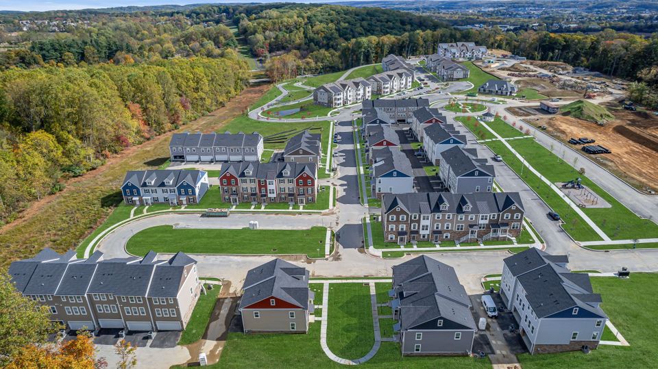 Aerial view of a suburban neighborhood with rows of townhouses and green lawns, surrounded by dense trees and distant hills.