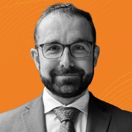A man with glasses, a beard, and a suit smiles against an orange background.