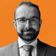 A man with glasses, a beard, and a suit smiles against an orange background.