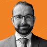 A man with glasses, a beard, and a suit smiles against an orange background.