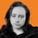 A man with long dark hair and a neutral expression poses in front of an orange background.