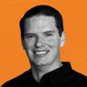 Smiling man with short dark hair wearing a black shirt against an orange background.