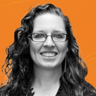 A woman with curly hair and glasses smiles against an orange background with subtle wave patterns.