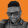 Man with styled hair, geometric designs on the side, wearing black-rimmed glasses and a button-up shirt, set against an orange background.