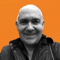A man with a shaved head smiles directly at the camera against an orange background, wearing a dark quilted jacket.