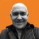 A man with a shaved head smiles directly at the camera against an orange background, wearing a dark quilted jacket.