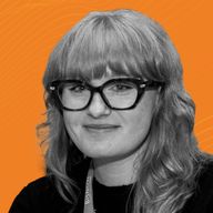A woman with glasses and curly hair poses against an orange background with subtle wave patterns.