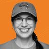 Smiling woman wearing glasses and a hat with a crab design, against an orange background.