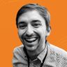 Smiling man with short hair and a checkered shirt, set against an orange background.