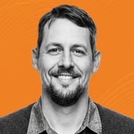 Smiling man with short hair and a beard, wearing a button-up shirt and sweater, set against an orange background with subtle wave patterns.