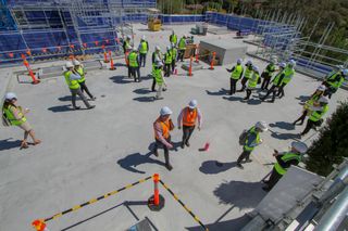 A group of people in hard hats and safety vests walk around a construction site with scaffolding and orange cones.
