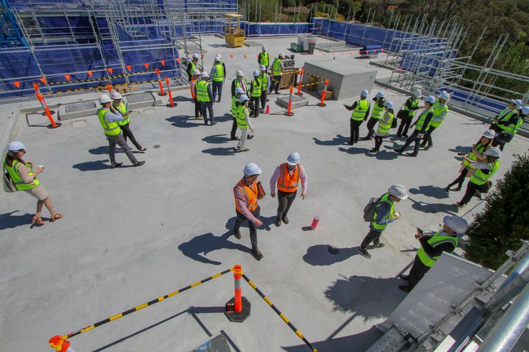 A group of people in hard hats and safety vests walk around a construction site with scaffolding and orange cones.