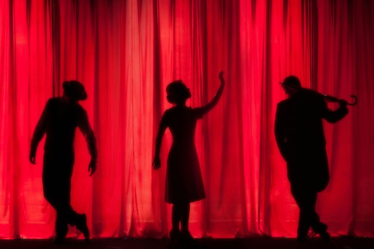 Silhouettes of three people striking poses in front of red stage curtains, with one holding an umbrella.
