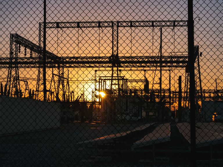 A power substation silhouetted against an orange sunset, viewed through a chain-link fence.