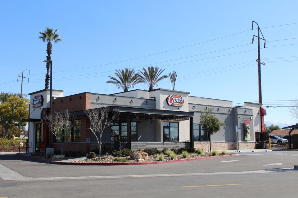 A Raising Cane's restaurant with palm trees, modern design, and outdoor seating. Clear blue sky in the background.