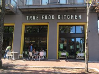 Entrance of True Food Kitchen with outdoor seating, featuring a group of people dining and trees lining the sidewalk.
